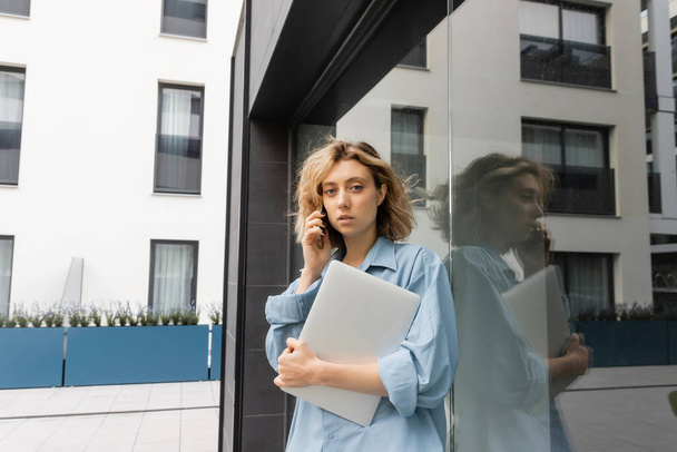 blonde woman with wavy hair talking on smartphone and holding laptop near hotel building in Barcelona  - Fotografie, Obrázek