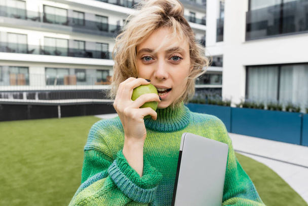 Blondine im Pullover hält Laptop in der Hand und isst grünen Apfel in der Nähe eines Hotelgebäudes in Barcelona  - Foto, Bild