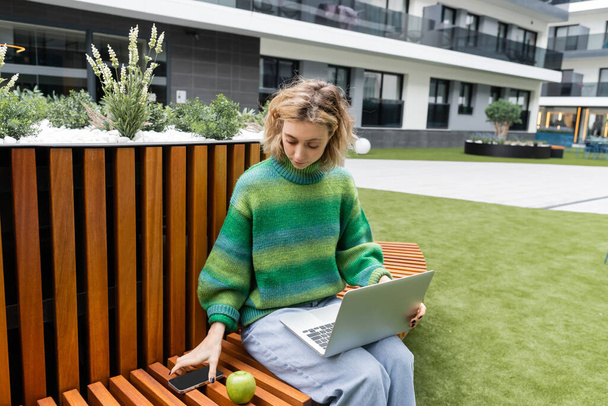 バルセロナのホテルの近くでスマートフォンを取りながらノートパソコンと木製のベンチに座っている緑のセーターのブロンドの女性  - 写真・画像