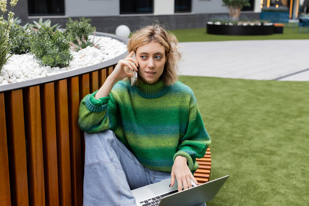 joven rubia en jersey verde sentada en banco de madera con portátil mientras habla en smartphone en Barcelona  - Foto, imagen