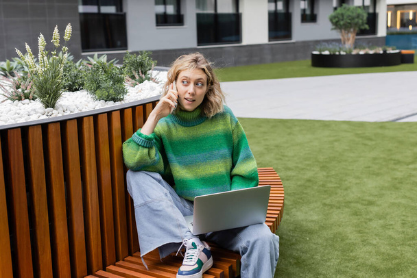 возбужденная молодая женщина в зеленом свитере сидит на деревянной скамейке с ноутбуком во время разговора на смартфоне  - Фото, изображение