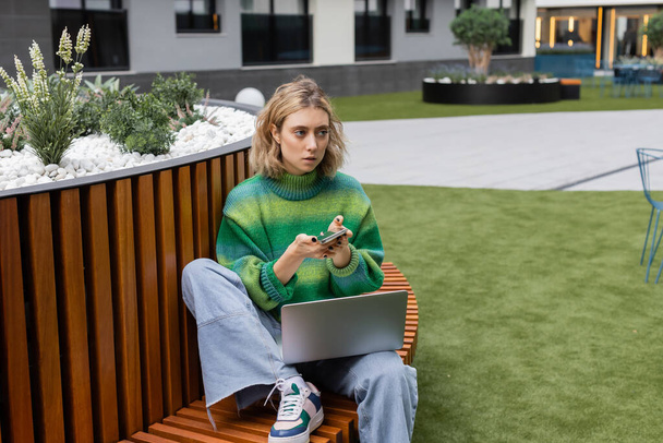 joven freelancer en jersey verde sentado en un banco moderno con portátil mientras usa el teléfono móvil  - Foto, Imagen
