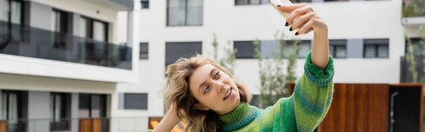 Счастливая женщина в свитере делает селфи на смартфоне возле отеля в Барселоне, баннер  - Фото, изображение