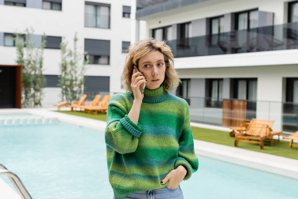 young woman in sweater standing with hand in pocket while talking on smartphone near outdoor swimming pool  - Fotografie, Obrázek