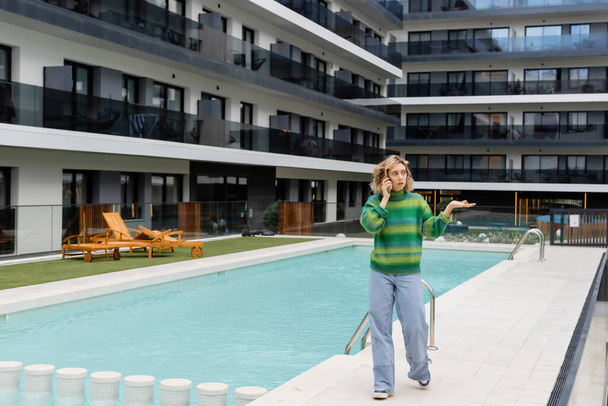 longitud completa de la mujer disgustada en suéter hablando en el teléfono inteligente cerca de la piscina al aire libre en Barcelona - Foto, imagen