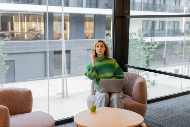 fiatal nő pulóverben beszél okostelefonon, miközben ül a laptop a szálloda halljában Barcelonában  - Fotó, kép