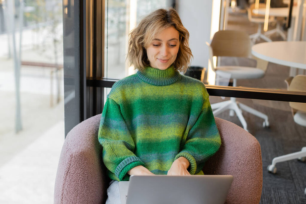 cheerful young woman in sweater using laptop in lobby of hotel in Barcelona - Φωτογραφία, εικόνα