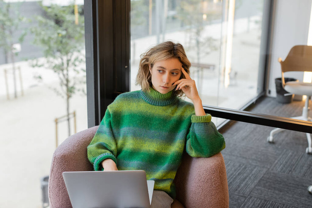 вдумчивая молодая женщина в свитере сидит с ноутбуком в вестибюле гостиницы в Барселоне - Фото, изображение