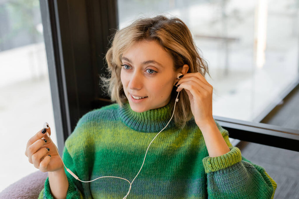 Lächelnde junge Frau in grünem Pullover mit kabelgebundenen Kopfhörern, während sie in der Lobby eines Hotels in Barcelona sitzt  - Foto, Bild