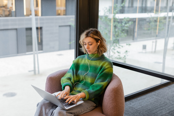 giovane freelance in maglione e auricolari cablati con computer portatile mentre è seduto nella hall dell'hotel a Barcellona  - Foto, immagini