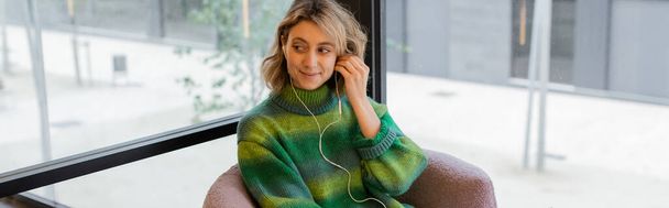 Весела жінка в зеленому светрі регулює навушники, сидячи в коридорі готелю в Барселоні, банер.  - Фото, зображення