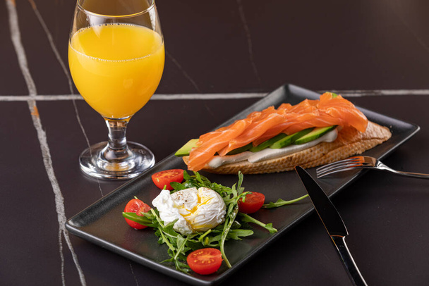 brxchetta with avocado and salted salmon. On a plate with poached egg. In the frame of a glass with orange juice and a plate with bruscheta. black background. Close-up. - Foto, Imagem