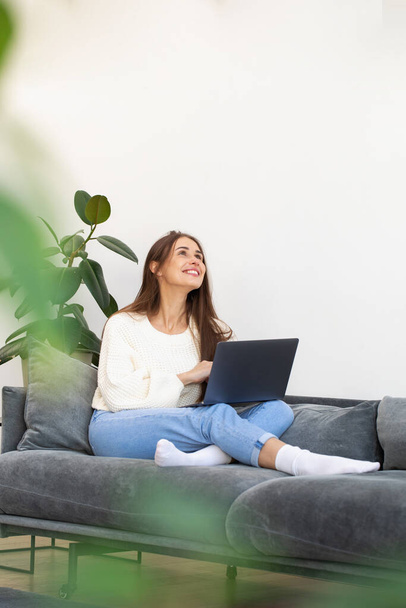A happy beautiful brunette enjoys shopping online, sitting on a sofa with a laptop on her lap. The concept of online shopping, using a laptop. - Fotoğraf, Görsel