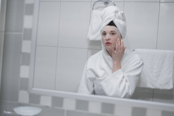 Woman reflected in mirror after morning shower with towel on head applies day facial cream close up, prevent first wrinkles, caring about skin use moisturizer creme, beauty treatment, skincare concept - Foto, Imagem