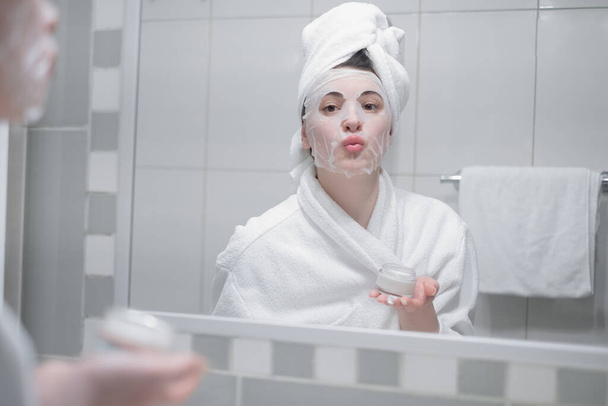 Woman reflected in mirror after morning shower with towel on head applies day facial cream close up, prevent first wrinkles, caring about skin use moisturizer creme, beauty treatment, skincare concept - Photo, Image
