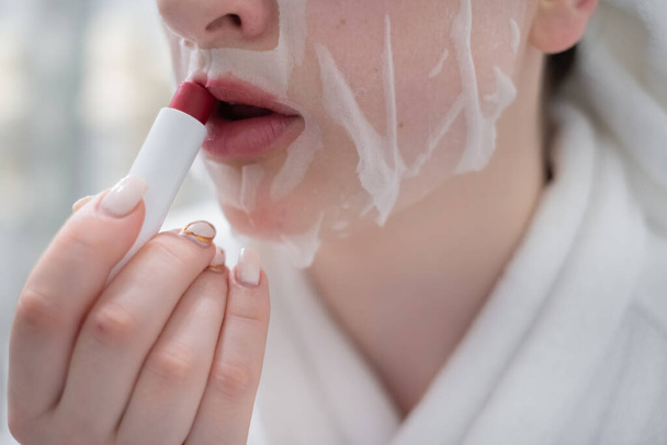 close-up of a woman holds a moisturizing lip mask, applies a cosmetic product on dry lips. Smiling women doing daily facials or healthy skin care, using scrub or balm. Body care concept. - Фото, изображение