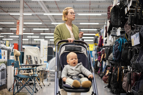 Casualy dressed mother choosing sporty shoes and clothes products in sports department of supermarket store with her infant baby boy child in stroller - Foto, immagini