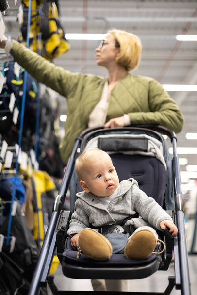 Rento pukeutunut äiti valitsemalla urheilullinen kengät ja vaatteet tuotteet urheilu osasto supermarket tallentaa hänen pikkulapsi poika lapsi rattaissa - Valokuva, kuva