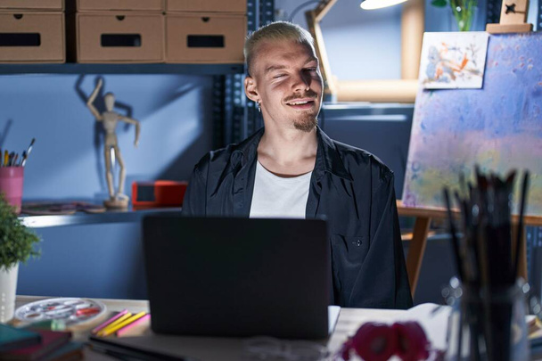 Young caucasian man using laptop at night at art studio winking looking at the camera with sexy expression, cheerful and happy face.  - Фото, зображення