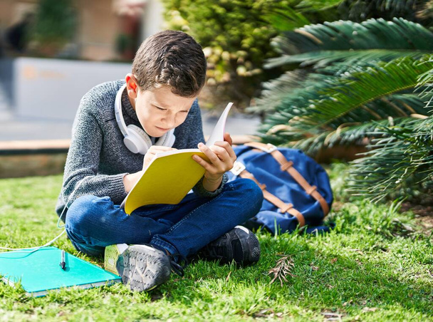 Блондин ребенок студент читает книгу сидя на траве в парке - Фото, изображение