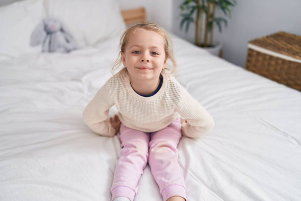 Αξιολάτρευτο ξανθό κορίτσι χαμογελά αυτοπεποίθηση κάθεται στο κρεβάτι στο υπνοδωμάτιο - Φωτογραφία, εικόνα