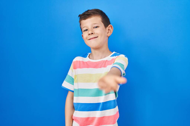 Junges kaukasisches Kind, das vor blauem Hintergrund steht und fröhlich lächelt und Handflächen anbietet, die Hilfe und Akzeptanz geben.  - Foto, Bild