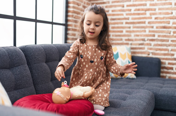 Entzückende hispanische Mädchen spielen mit Baby-Puppe sitzt auf Sofa zu Hause - Foto, Bild