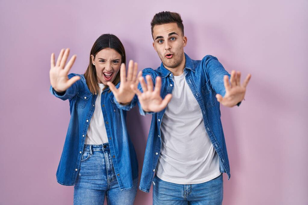 Young hispanic couple standing over pink background doing stop gesture with hands palms, angry and frustration expression  - Photo, Image