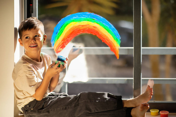 Happy small european boy chooses paints, draws rainbow on window in children room interior. Elementary education, entertainment and fun, dreams, art and fantasy, childhood at home and children garden - Photo, Image