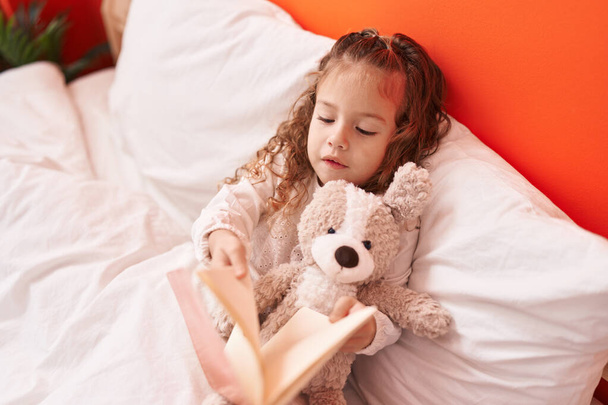Adorable rubia niño leyendo libro acostado en la cama con oso de peluche en el dormitorio - Foto, imagen