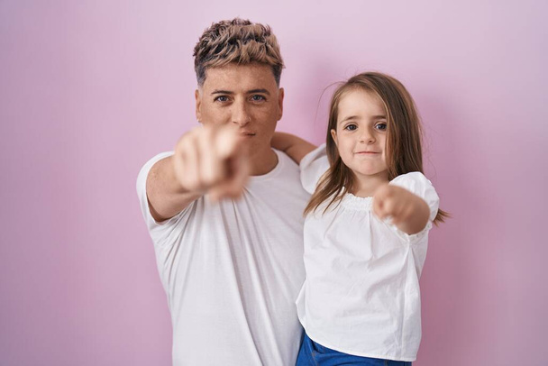 Junger Vater umarmt Tochter vor rosa Hintergrund und zeigt mit dem Finger in die Kamera und zu dir, selbstbewusste Geste, die ernst wirkt  - Foto, Bild
