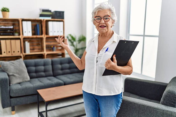 Oudere grijs-harige vrouw psycholoog houdt klembord bij psychologie centrum - Foto, afbeelding