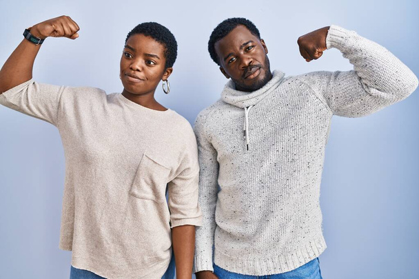 Young african american couple standing over blue background together strong person showing arm muscle, confident and proud of power  - Photo, Image