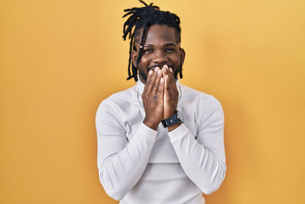 Afrikaanse man met dreadlocks dragen coltrui over gele achtergrond lachen en beschaamd giechelen bedekken mond met handen, roddel en schandaal concept  - Foto, afbeelding