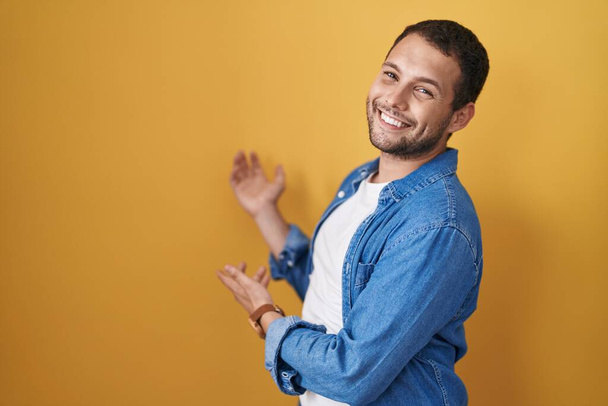 Spaanse man die over gele achtergrond staat en uitnodigt om met open hand lachend binnen te gaan  - Foto, afbeelding