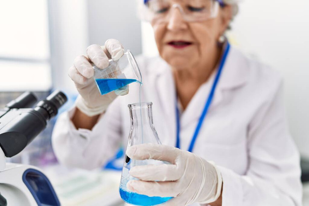 Senior grey-haired woman wearing scientist uniform pouring liquid at laboratory - Photo, Image
