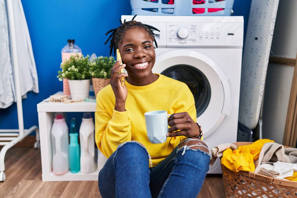 Femme afro-américaine parlant sur le smartphone buvant du café en attendant la machine à laver à la buanderie - Photo, image