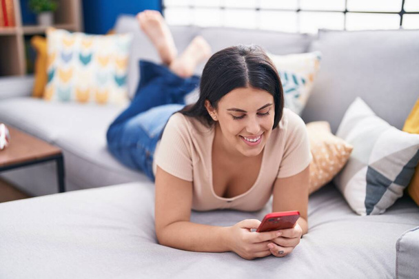 Nuori kaunis latino nainen käyttää älypuhelinta makaa sohvalla kotona - Valokuva, kuva