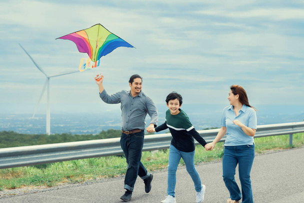 Progressive happy carefree family vacation concept. Young parents mother father and son run along and flying kite together on road with natural scenic on mountain and wind turbine background. - Photo, Image