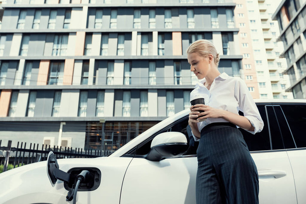 Businesswoman drinking coffee, leaning on electric vehicle recharging at public charging station with residential apartment condos building in background as progressive lifestyle by eco-friendly car. - Photo, image