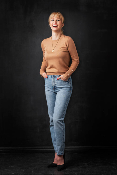 Full length of attractive blond haired woman standing at isolated dark background. Happy female wearing sweater and jeans and looking at camera and smiling. Copy space. - Foto, imagen