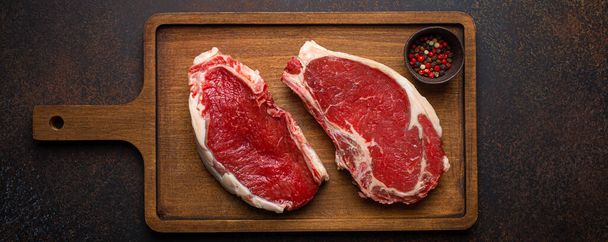 Zwei rohe Rindfleisch Rib Eye marmorierte Steaks auf hölzernem Schneidebrett mit Gewürzen auf dunklem rustikalem Hintergrund, bereit zum Grillen von oben, Zubereitung des Abendessens mit Fleisch - Foto, Bild