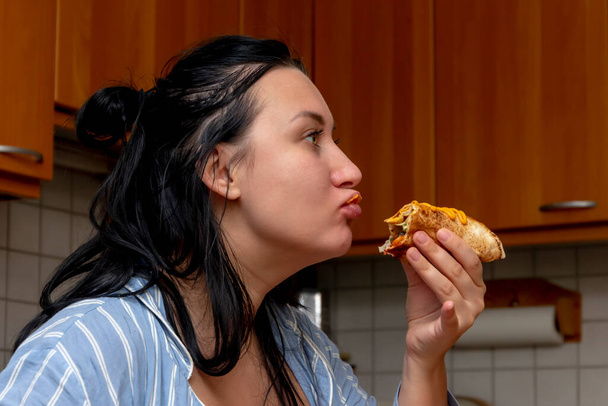 A plump young woman eats pita bread with meat on the background of the kitchen. Concept: passion and attachment to food, diet and fasting. - Foto, Imagem
