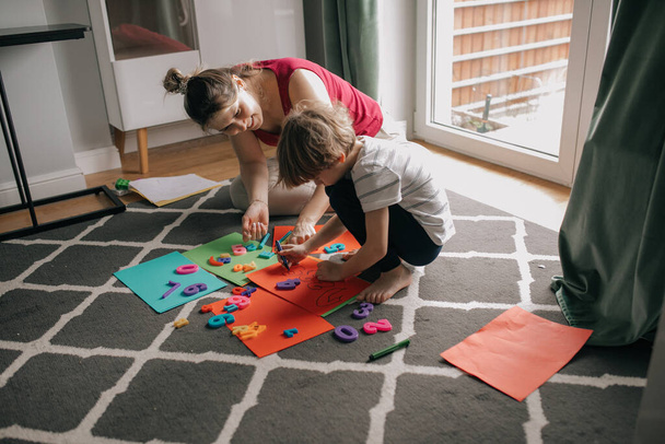 Happy mother and little son play together sitting on carpet at home room. Draw markers on colored paper sheets, learning numbers, numeration. Educational games, preschooler, home study. - Foto, Imagem