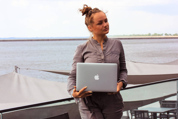 Kyiv, Ukraine. May 1, 2021 A young woman in business suit is standing against the sea, holding an Apple laptop. Serious girl working remotely on cafe restaurant hotel resort terrace Hipster lifestyle. - Fotografie, Obrázek