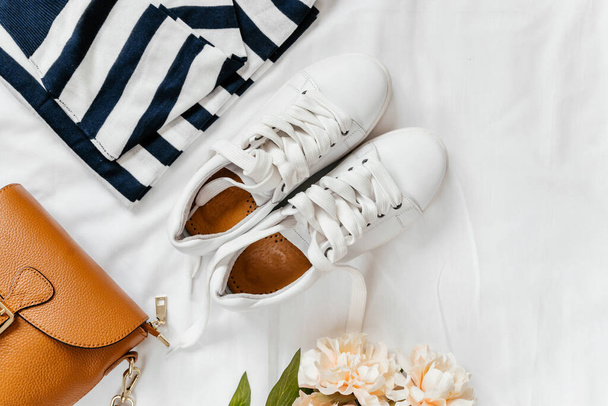 Blue jeans, t shirt, bag and white sneakers on white bed. Trendy clothes. Women's casual spring summer outfit. Fashion concept. Flat lay, top view - Fotó, kép