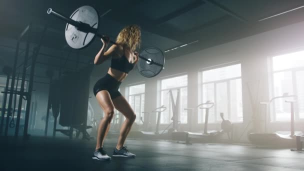 Close-up shot of a confident girl with curly, blonde hair doing squats with a barbell while exercising in the gym. Active, fit lady testing her strength with intensive workout. High quality 4k footage - Video