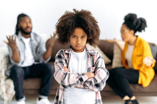 Difficulties in the family. An African American unhappy preschool girl looks offendedly at the camera, against the background, parents sort things out, shout at each other, swear, the child is scared - Foto, Imagem