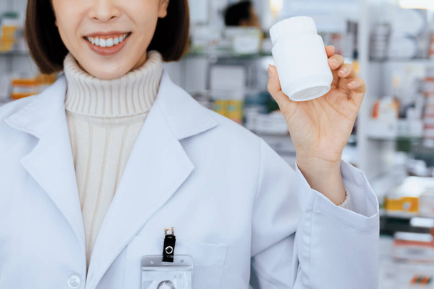 Closeup fiatal affable gyógyszerész show qualified medicine pill palack mockup termék copyspace a gyógyszertárban. Gyógyszerész a naplójában dolgozik a gyógyszertárban, gyógyszeres doboz a polcon a háttérben. - Fotó, kép