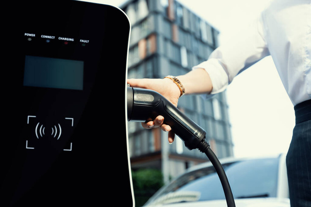 Focus EV charger plug and electric car at public charging station with blur progressive businesswoman holding charger and apartment condo building in background. Eco friendly electric vehicle concept. - Foto, imagen